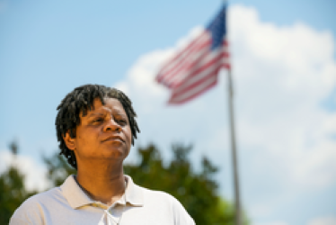 Person looking up and past camera, with American flag waving in wind far behind them, against blue sky.