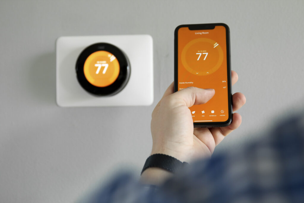 Mobile phone with smart home app next to a smart thermostat, showing temp of 77 degrees on orange background.